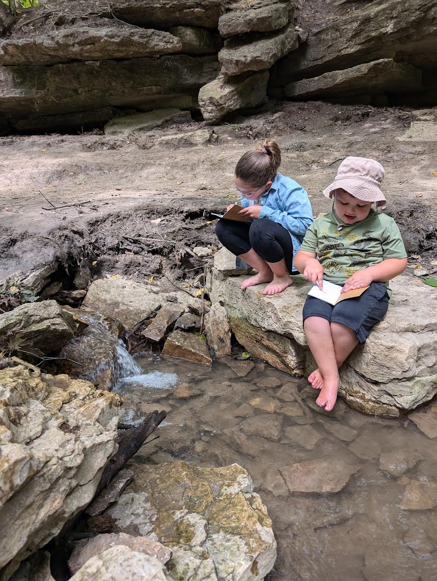 two children draw in notebooks near water